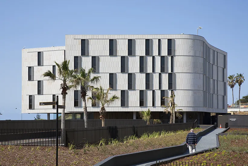 Modern building with a wavy design and a silver facade, surrounded by palm trees. A person walks on a pathway leading to the entrance. Bright blue sky in the background.