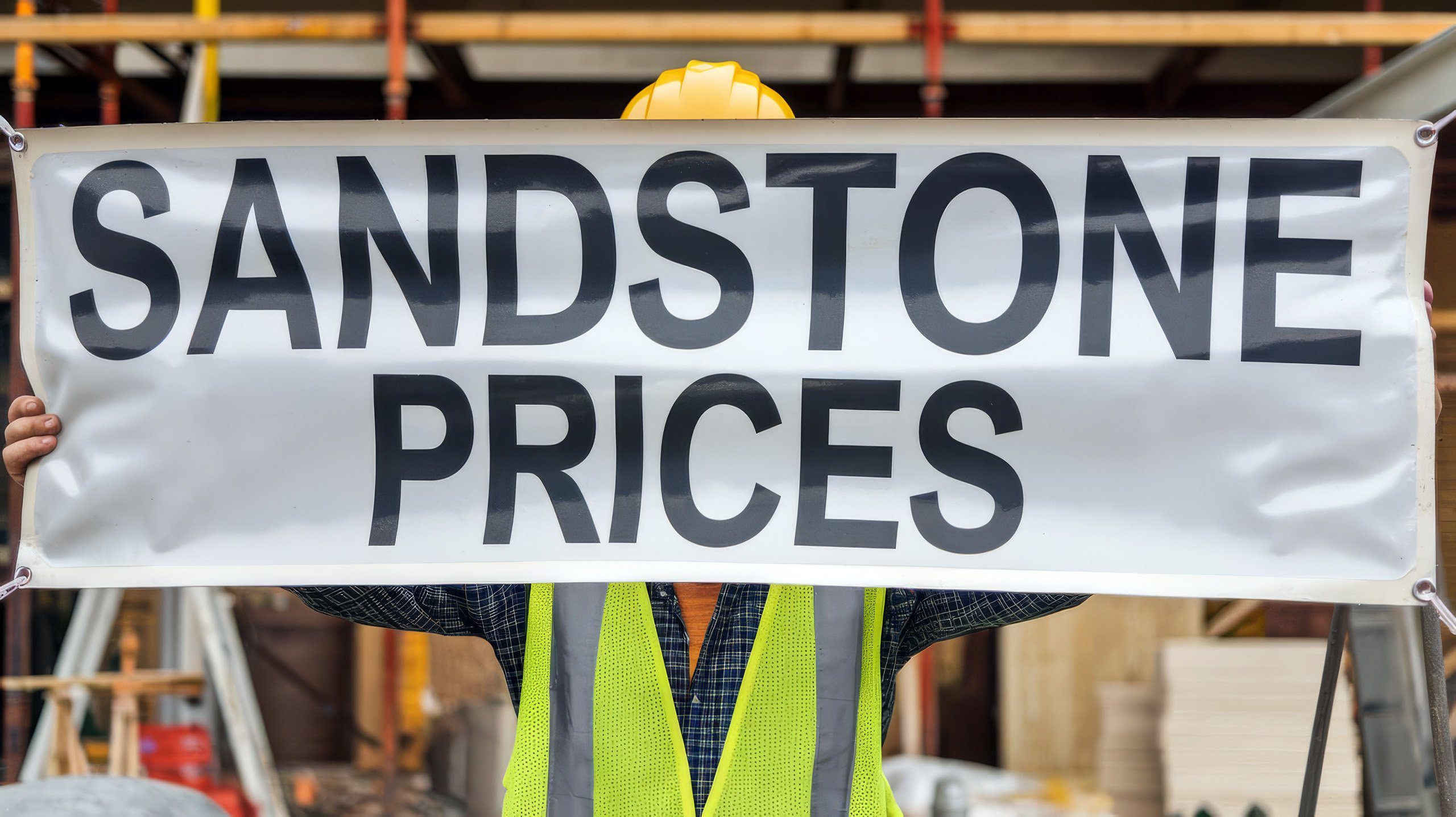 A person in a construction vest and hard hat holds a banner that reads "SANDSTONE PRICES" in large, bold letters. They stand in front of a construction site with scaffolding and building materials in the background.
