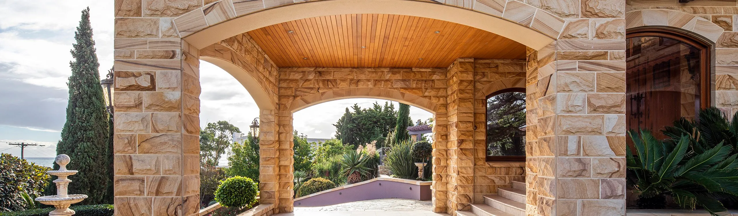 Stone archway entrance of a luxurious house, with a wooden ceiling and large stone columns. The view includes a paved path, greenery, and a glimpse of a fountain on the left, with a clear sky in the background.