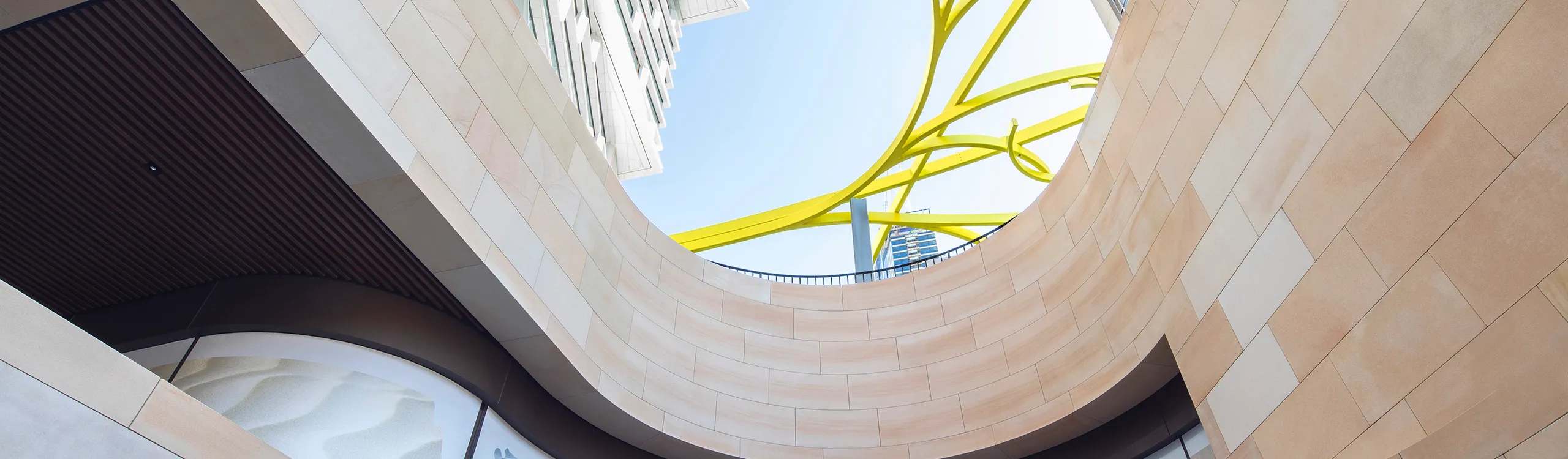 View from the ground looking up through a circular architectural opening surrounded by modern beige buildings, with bright yellow abstract sculpture elements and a clear blue sky above.