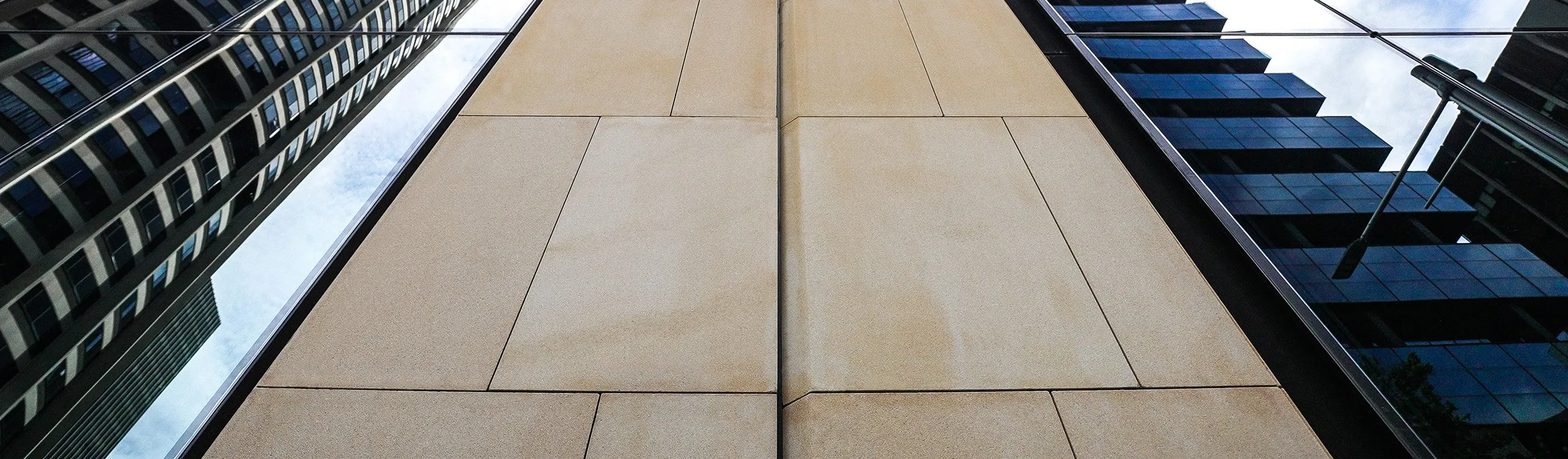 Upward view of a modern building facade, with glass and stone sections reflecting the sky and adjacent structures. The composition creates symmetry, highlighting geometric shapes and contrasting textures.