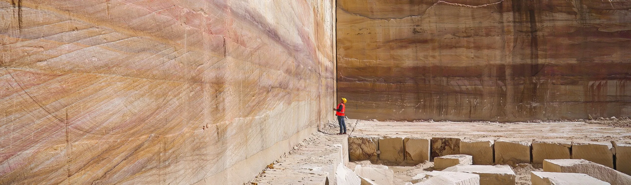 A person in a red shirt and yellow helmet stands against a massive sandstone wall in a quarry. The wall displays natural layers of hues, while large stone blocks are scattered on the sandy ground.