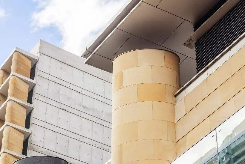 Modern building exterior featuring round and rectangular beige-tiled columns, concrete walls, and metal overhangs against a partly cloudy sky. The photo highlights architectural lines and geometric shapes. Modern building exterior featuring round and rectangular beige-tiled columns, concrete walls, and metal overhangs against a partly cloudy sky. The photo highlights architectural lines and geometric shapes.
